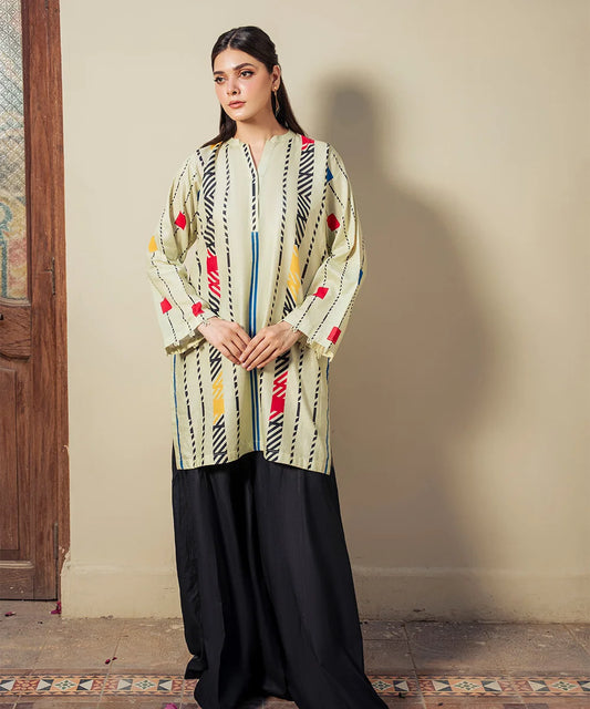Woman wearing a patterned top and black pants standing against a neutral wall.